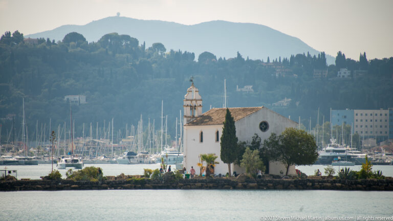 White Church of Panagia Vlacherna on a small islet, with bell tower, cypress trees, marina masts, and wooded hills behind.