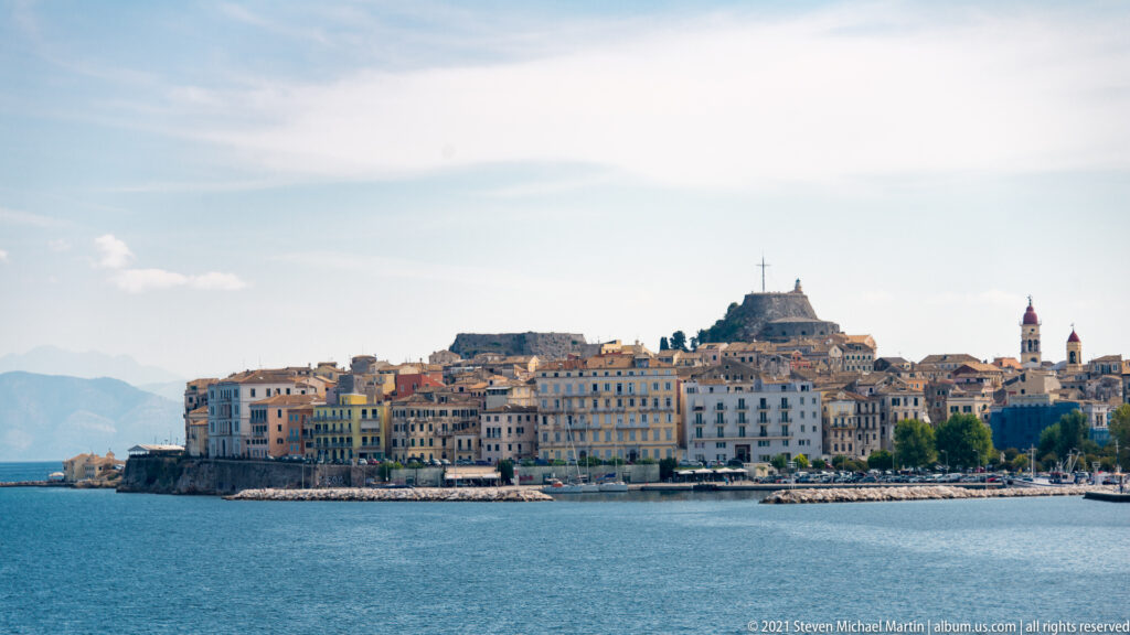 Corfu Town skyline across calm blue water, with pastel waterfront buildings, fortified heights, and church towers.
