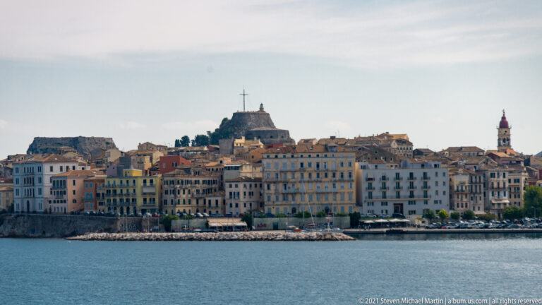 Historic Corfu Town buildings along the waterfront beneath a fortified hill, with church tower and cross visible above the roofs.
