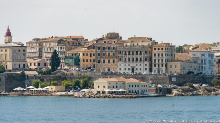 Historic Corfu waterfront with closely packed multistory buildings above rocky shore and low seafront structures.