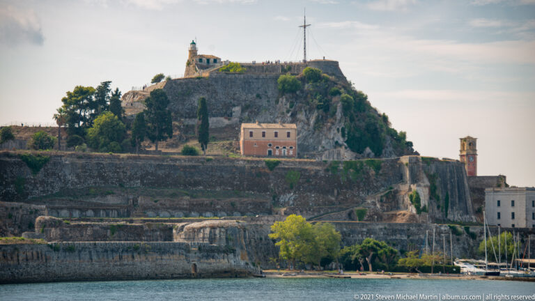 Seaward view of Corfu's Old Fortress with rocky summit, layered stone walls, upper buildings, and a bell tower near the water.