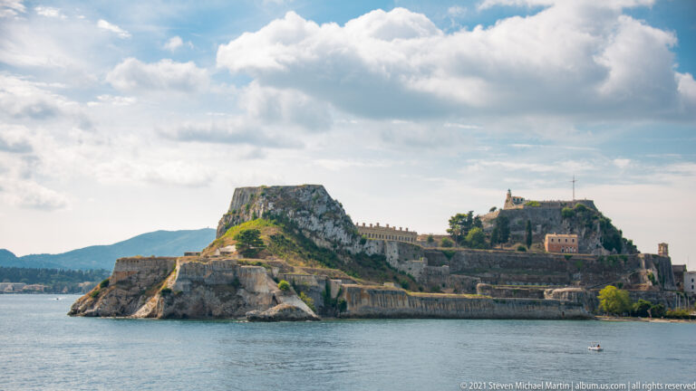 Wide seaward view of Corfu's Old Fortress with twin rocky heights, layered bastions, sea walls, vegetation, and clouds.