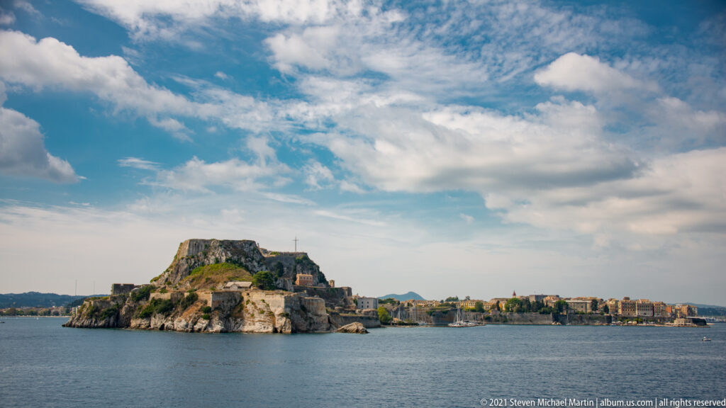 Wide seaward view of Corfu's Old Fortress on a rocky promontory, with Corfu Town extending along the distant shoreline.