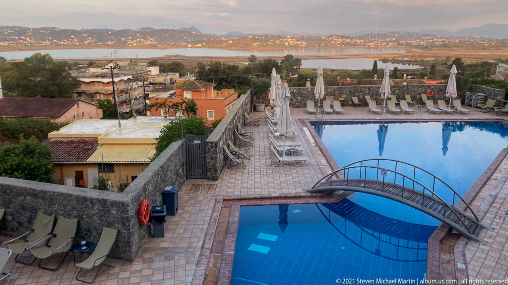 Elevated outdoor swimming pools with arched footbridge, lounge chairs, umbrellas, stone terrace, and distant Corfu coastline.