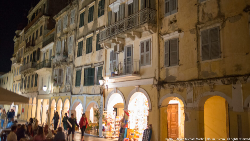 Night street in Corfu Town with pedestrians, lit shops under arcades, shuttered windows, balconies, and weathered façades.