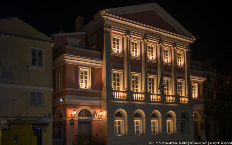 Illuminated Ionian Bank Building with pink neoclassical façade, white pilasters, pediment, and arched stone ground floor.