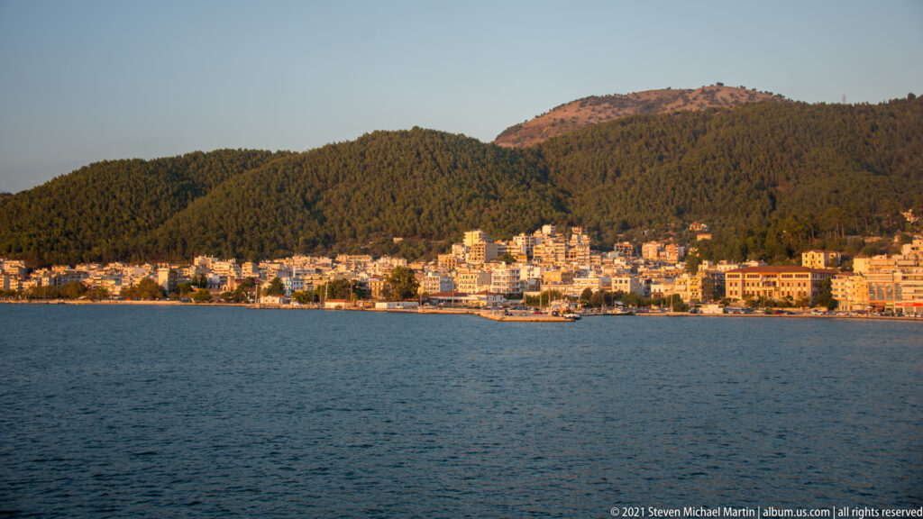 Waterfront buildings along Corfu shoreline beneath densely forested hills, viewed across open water.