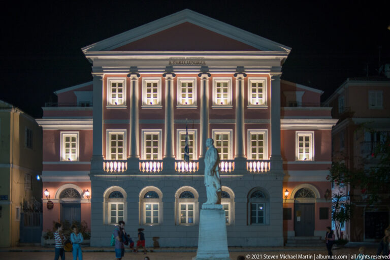 Statue of Georgios Theotokis before the illuminated pink-and-white Ionian Bank Building on Heroes of Cypriot Struggle Square.