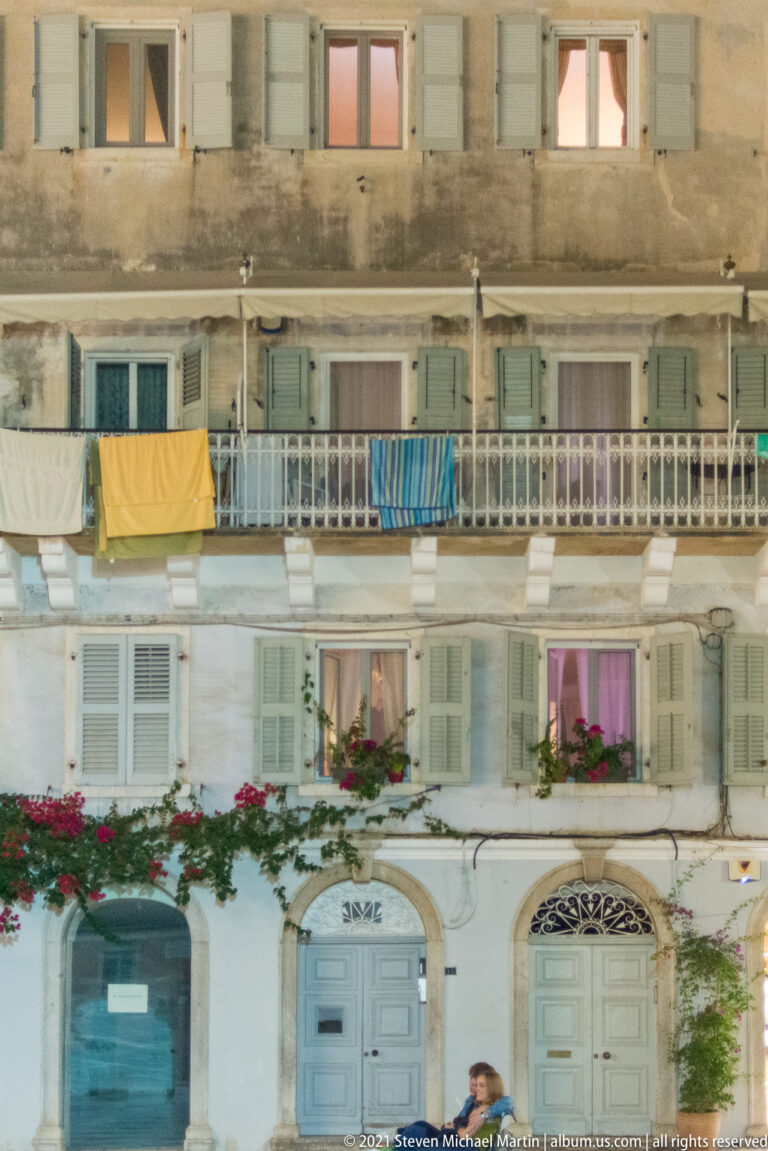Weathered multi-story façade with pale blue shutters and doors, iron balconies, laundry, and red flowers.