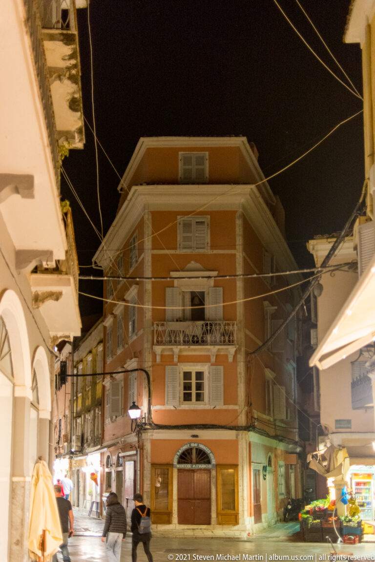 Narrow night street with overhead wires, pedestrians, arcaded shops, and an ochre corner building with shutters and balcony.