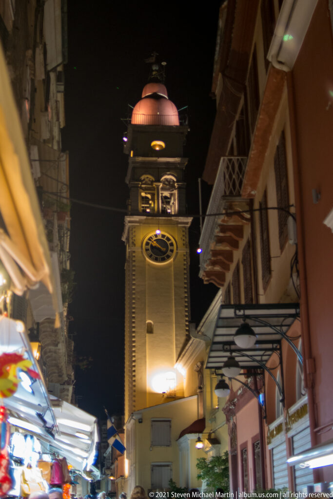 Illuminated bell tower of the Church of Saint Spyridon rising above shop awnings and balconies on a narrow Corfu street.