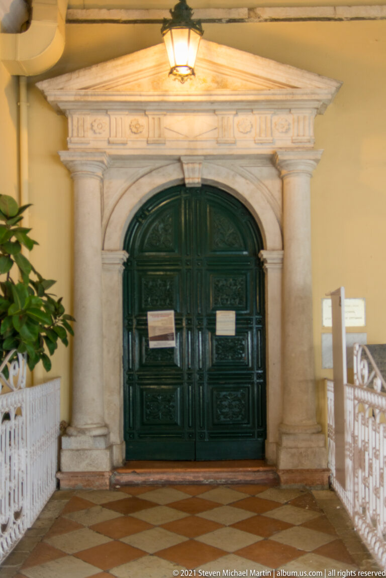 Dark green paneled double doors within a carved pale stone portal, lit by a hanging lantern above a patterned tile floor.