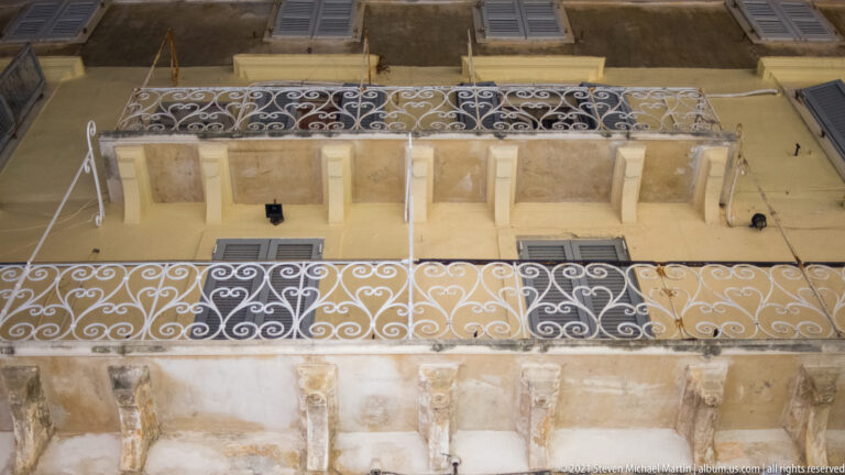 Two stacked white wrought-iron balconies with scrollwork on a weathered ochre façade with shuttered windows.