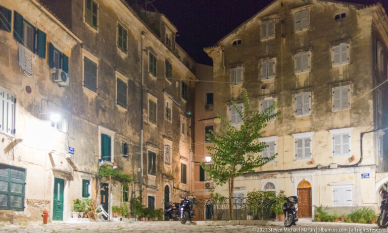 Small tree in a paved night courtyard surrounded by tall weathered façades with shutters, wiring, plants, and air-conditioning units.