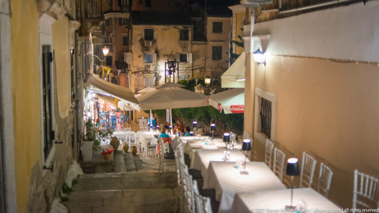 Restaurant tables and white chairs arranged along a stepped stone lane descending toward lit shops in Corfu Town at night.