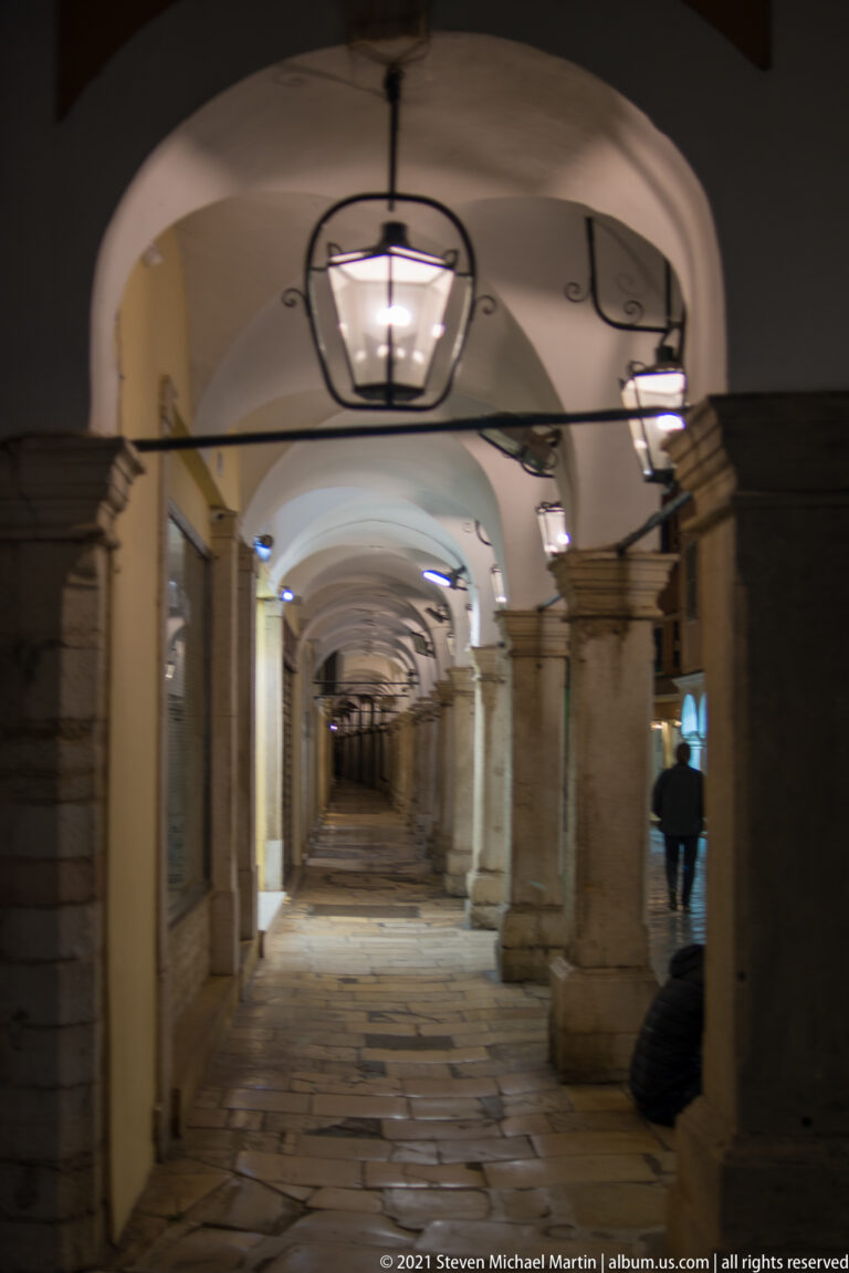 Long covered stone arcade with repeated columns, rounded arches, hanging lanterns, and worn paving receding into the distance.