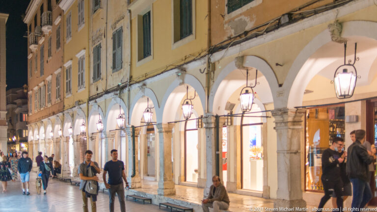 Bright shopfronts beneath a long arcade of pale arches and stone columns, with pedestrians and hanging lanterns at night.