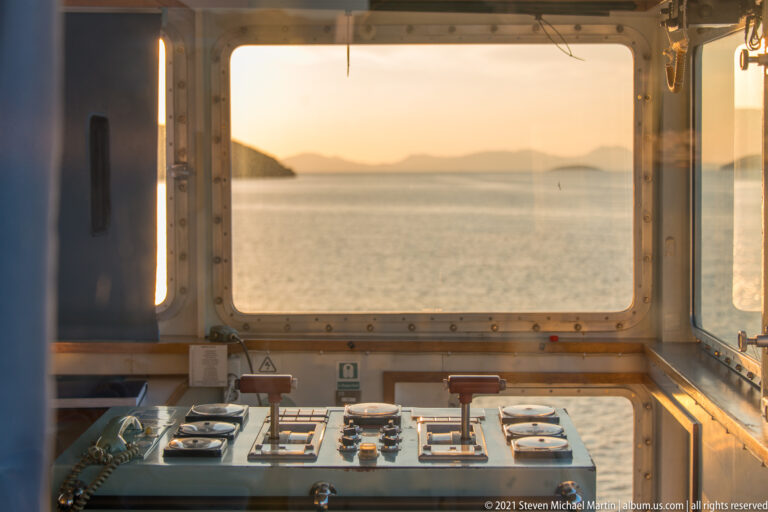 Ferry bridge console beneath wide windows overlooking calm sea and distant islands in warm evening light.