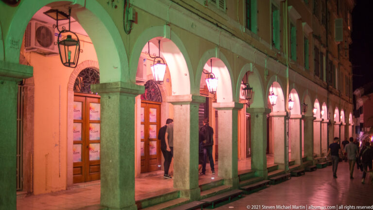 Long arcade at night washed in green and red light, with stone columns, rounded arches, hanging lanterns, and pedestrians.