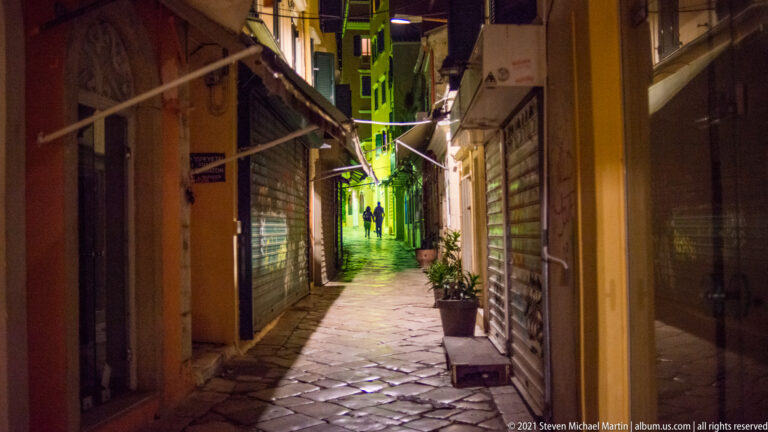 Narrow stone-paved Corfu lane at night with closed shops, overhead awnings, potted plants, green light, and two distant pedestrians.