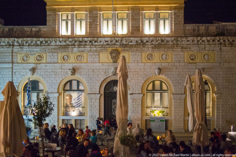 Nighttime café tables and diners before an illuminated historic façade with arched openings, rusticated masonry, and portrait medallions.