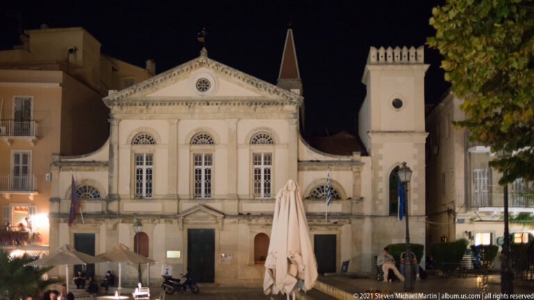 Illuminated pale historic building with triangular pediment, arched windows, square tower, café umbrellas, and scooters at night.