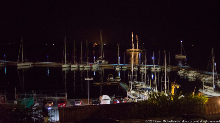 Moored sailboats and tall masts in a dark Corfu marina, with harbor lights reflected on the water and a breakwater beyond.