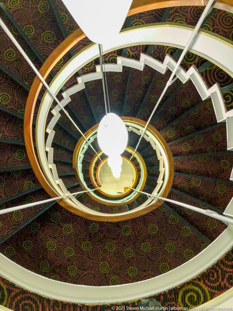 Upward view through a spiral stairwell with white curved rails, patterned ceiling panels, and suspended globe lights.