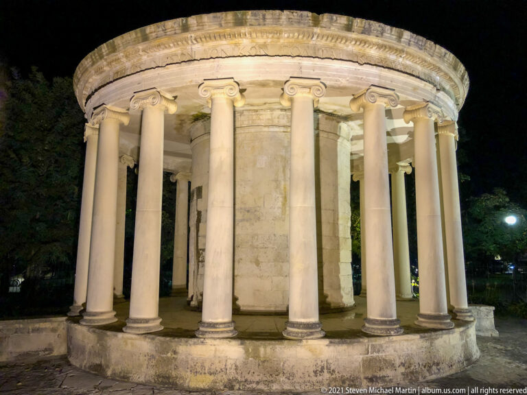Round pale-stone pavilion at night with a ring of slender columns, curved entablature, and dark trees behind.