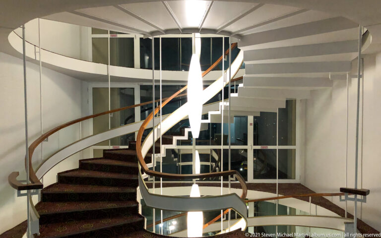 Curving white multistory staircase with dark wooden handrail, glazed walls, and bright vertical lighting in a modern interior.