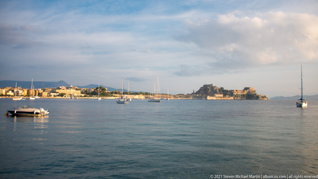 Corfu bay with anchored sailboats, waterfront buildings, and the rocky fortified mass of the Old Fortress in the distance.
