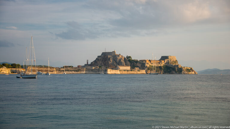 Old Fortress of Corfu across calm water, with an anchored sailboat at left and layered stone defenses on the rocky promontory.
