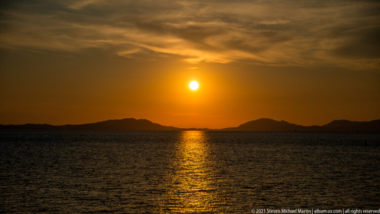 Orange sunset above silhouetted low islands, with a bright reflection extending across dark sea water.