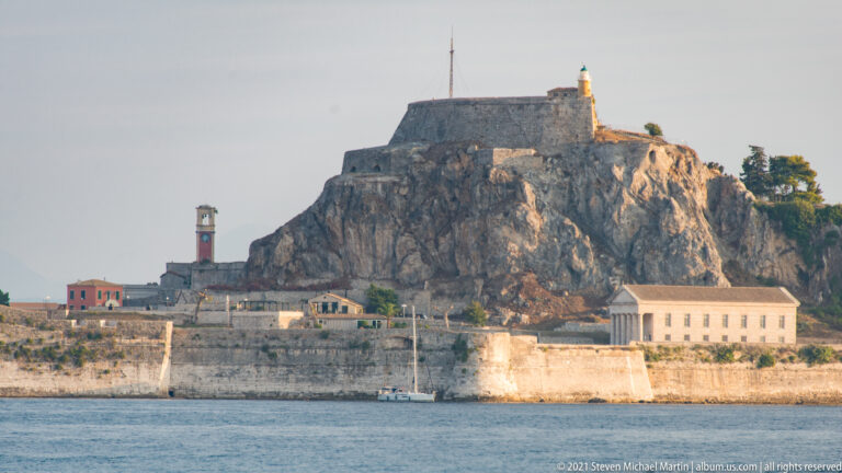 Close view of Corfu's Old Fortress with massive stone walls climbing a rocky summit and a lighthouse above waterfront buildings.