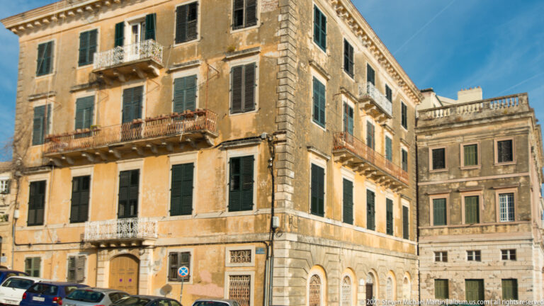 Weathered ocher corner building in Corfu with green shutters, balconies, stone trim, and an adjoining pale historic façade.