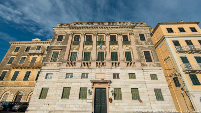Formal historic Corfu façade with pale stone base, green shutters, pilasters, central balcony, and balustraded roofline.