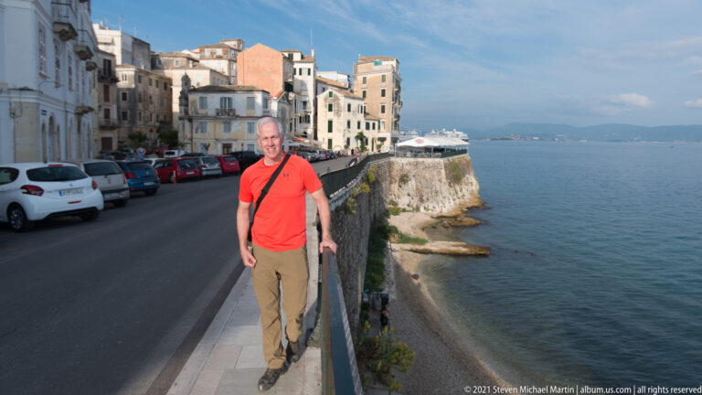 Person in red shirt standing beside a stone seawall on Corfu's waterfront, with parked cars, historic buildings, and sea beyond.