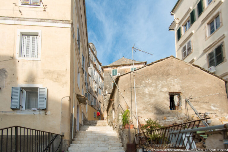 Stone steps climbing a narrow Corfu residential passage between weathered buildings with shutters, railings, antennas, and potted plants.