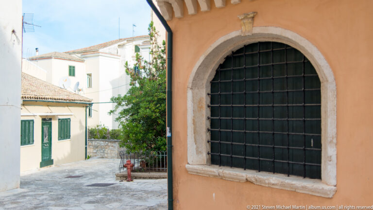 Arched window with pale stone surround and black iron grille in an ocher wall beside a small paved courtyard.