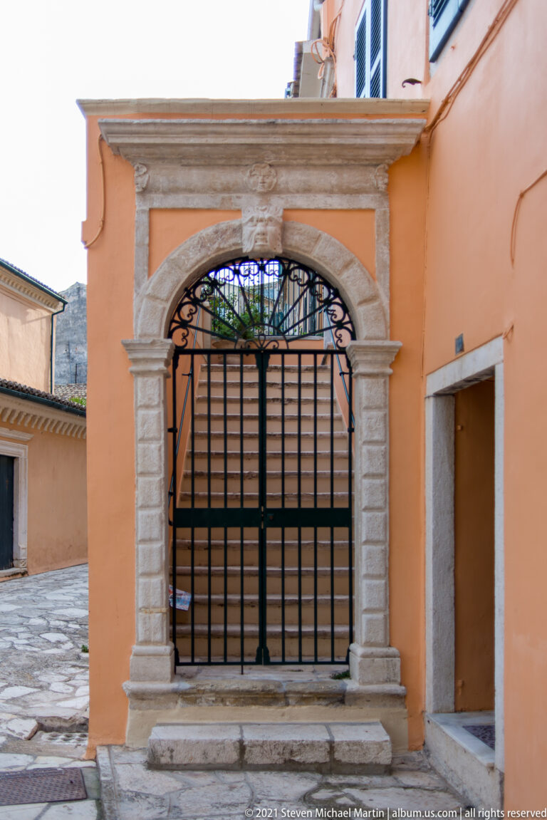 Large black metal grille within an ornate pale-stone arched surround and cornice set in an ocher courtyard wall.