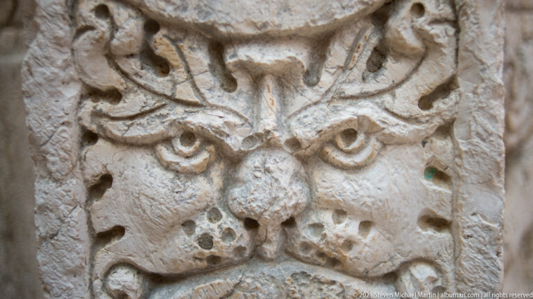 Weathered carved mask on the 1699 Kremasti Square cistern wellhead, with heavy brows, staring eyes, and curling ornament.