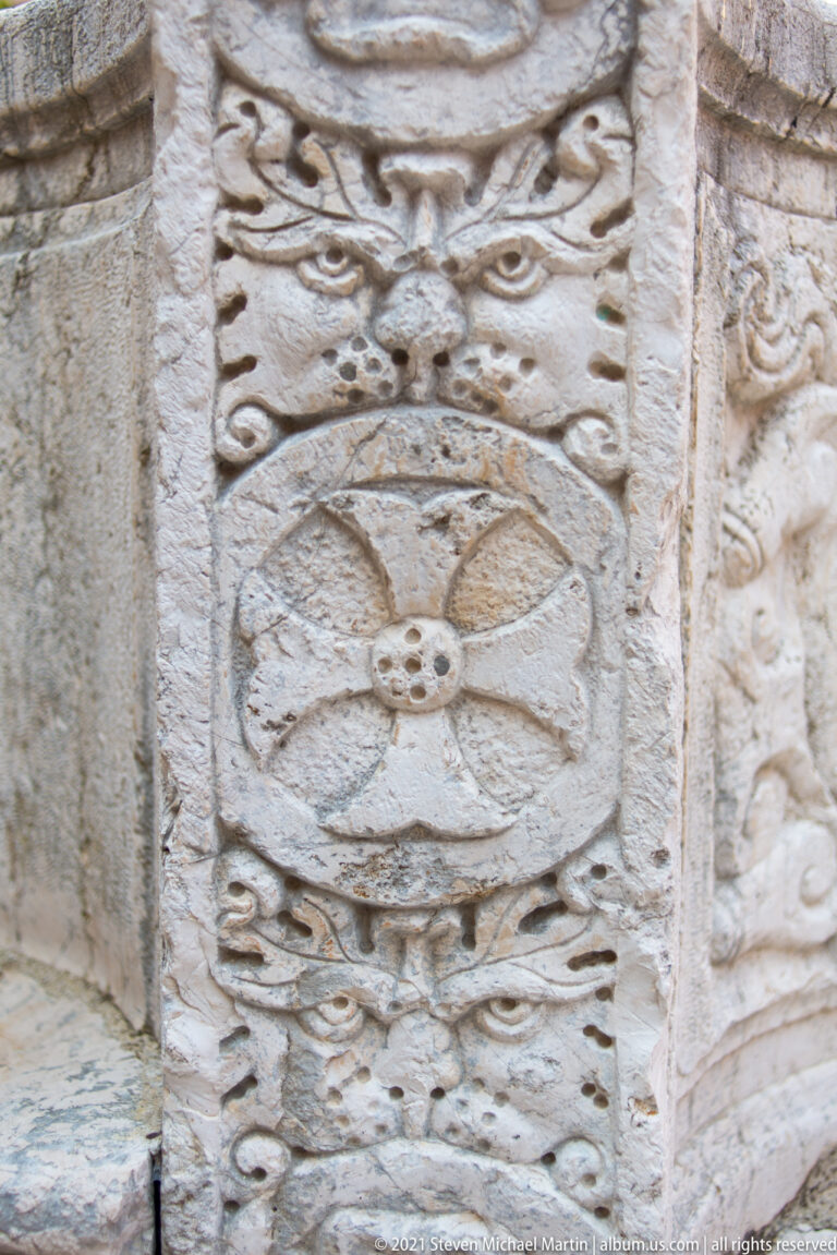 Carved rosette and mask reliefs on the weathered stone wellhead of the 1699 Kremasti Square cistern.