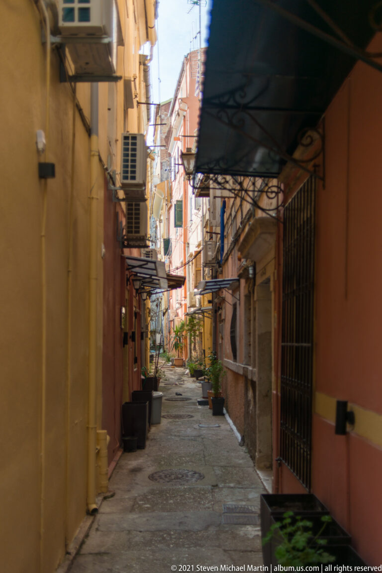 Narrow paved Corfu alley between warm-colored buildings with awnings, barred windows, air conditioners, balconies, and potted plants.