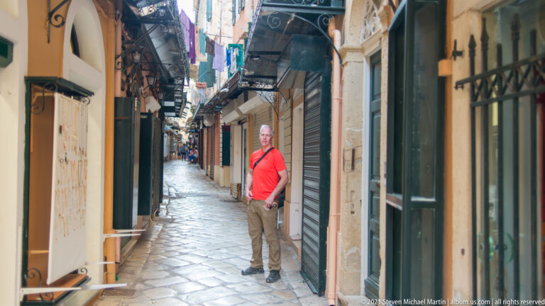 Person in red shirt standing in a narrow stone-paved Corfu lane lined with shuttered shops, signs, awnings, and balconies.