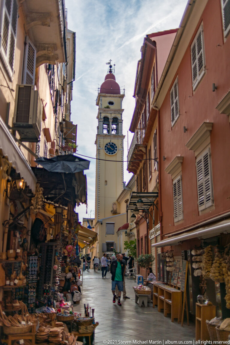 Corfu market lane lined with souvenir displays and awnings, with pedestrians and a pale bell tower topped by a red dome ahead.