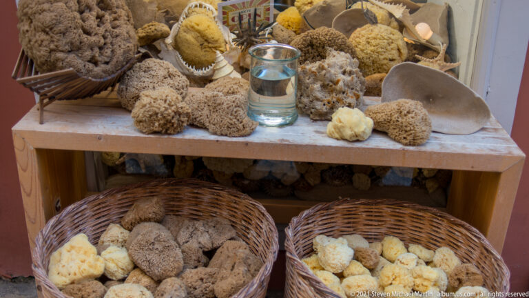 Assorted tan, cream, and gray natural sea sponges piled in wicker baskets and on a shop shelf around a glass vessel.