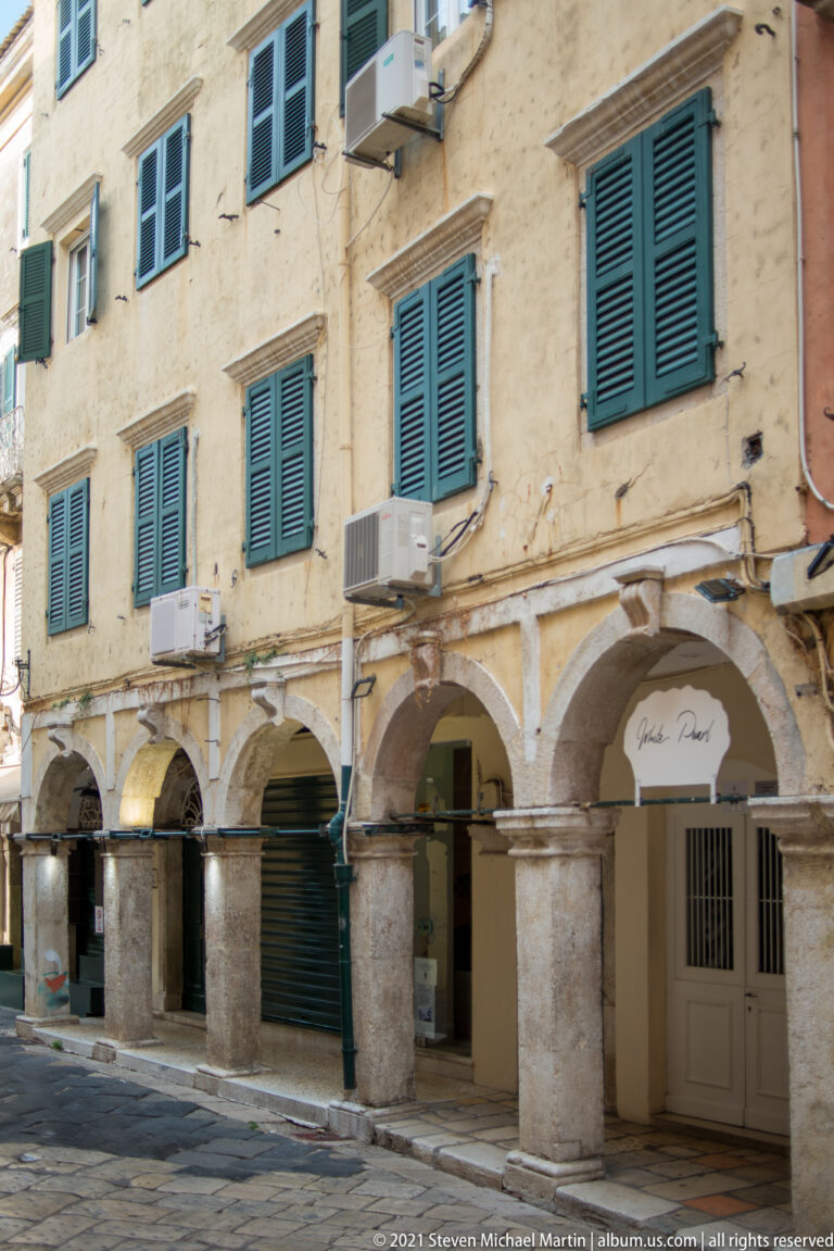 Weathered Corfu building with green shutters above a ground-floor stone arcade of repeated round arches and heavy masonry piers.