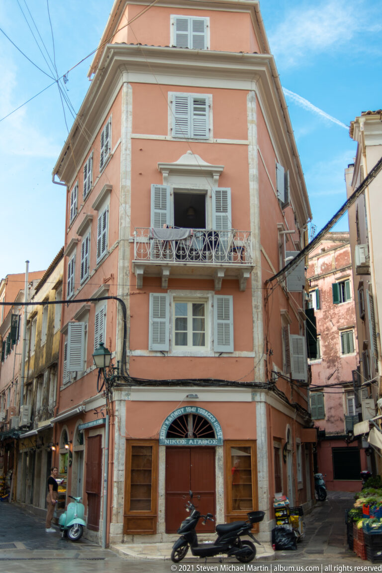 Salmon-colored Corfu corner building with pale shutters, stone trim, balcony, closed ground-floor shopfronts, and scooters at the intersection.