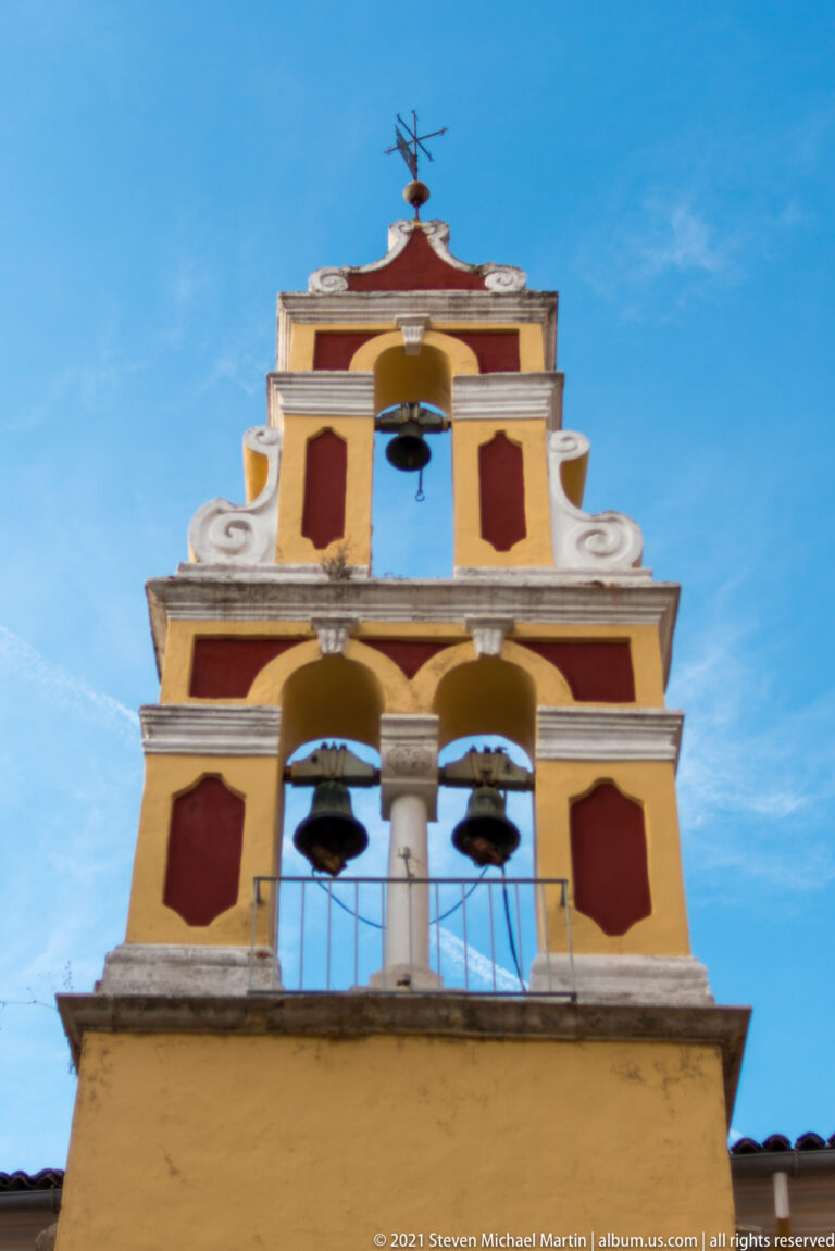 Low-angle view of a yellow bell tower with three dark bells, white scrollwork, red panels, and a weather vane against blue sky.