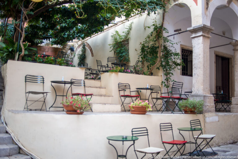 Terraced courtyard café with metal tables and chairs, pale arches, stone steps, climbing plants, and pots of flowers.